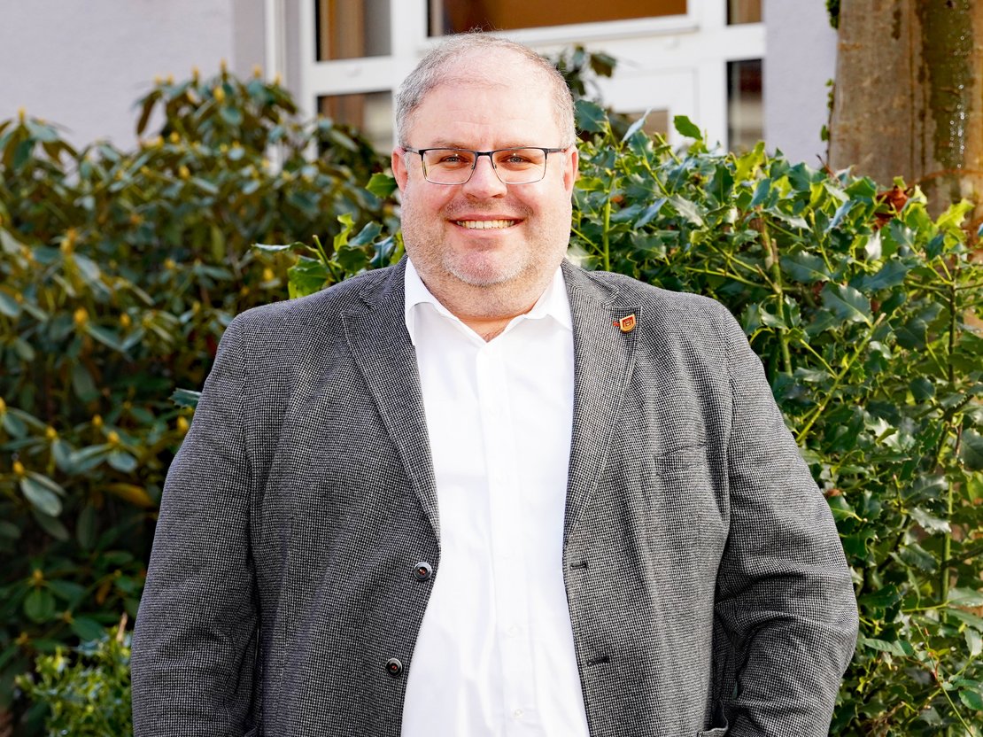 Fachbereichsleiter Hoch- und Tiefbau Sebastian Jülich vor dem Ratstrakt, vor grünen Büschen, lächelnd.