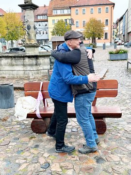 Die Bürgermeister Alexander Brodführer und Ulrich Schulte umarmen sich vor der Holzbank.