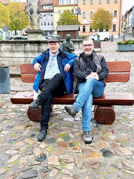 Die beiden Bürgermeister Ulrich Schulte und Alexander Brodführer sitzen eng beieinander auf einer Holzbank.
