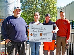 Bild von der Übergabe des Spendenschecks auf der Brücke an der Halle des städtischen Baubetriebshofs, vier Personen, davon eine Frau und drei Männer in Arbeitskleidung.