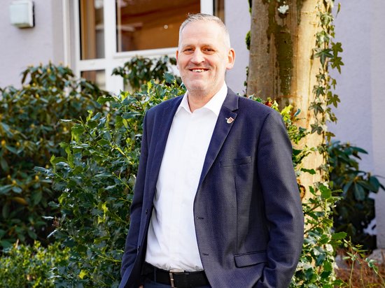Der allgemeine Vertreter Matthias Steinhoff vor dem Ratstrakt, vor grünen Büschen, lächelnd.