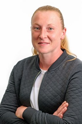 Portraitfoto von Evi Schmidt, lächelnd, starke Pose mit überschlagenen Armen, blond, lange Haare zum Zopf.