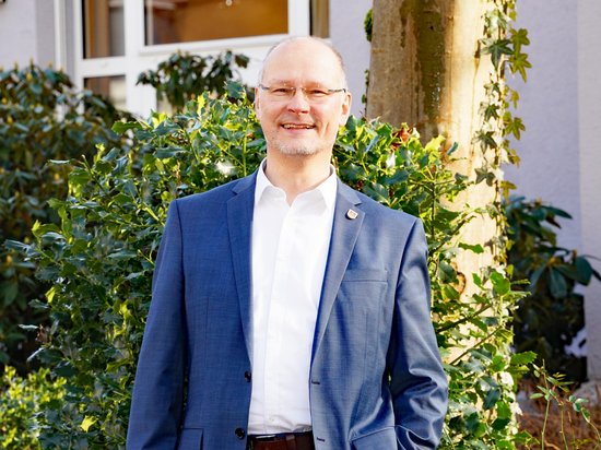 Bürgermeister Ulrich Schulte vor dem Ratstrakt, vor grünen Büschen, lächelnd.